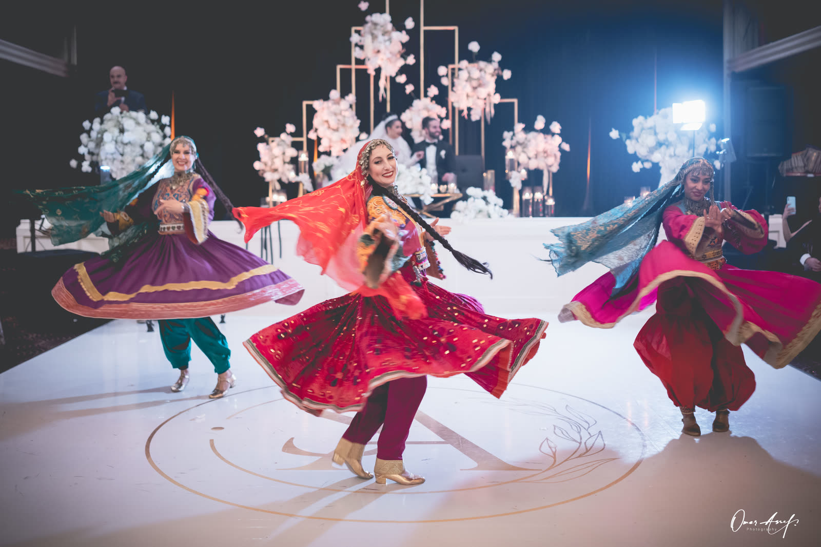 Three dancers in red, purple and pink Afghan dresses twirling in front of the bride and groom